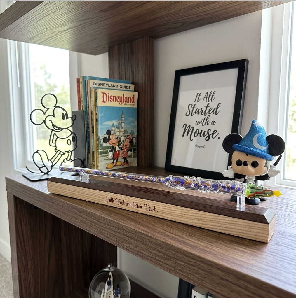 A personalised wooden keepsake wand stand engraved with "Faith, Trust and Pixie Dust" sits on a wooden shelf next to a Mickey silhouette card, Disneyland guidebooks, a framed quote print, and a Mickey figurine.
