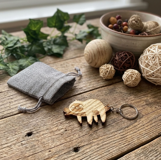 A wooden sloth keychain with a silver metal ring, displayed on a rustic wooden table alongside a small grey fabric pouch, decorative balls, and green leaves.