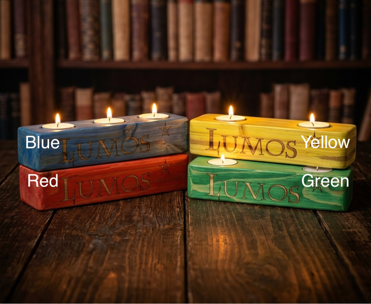 Four rectangular wooden candle holders with tea light candles, labelled Blue, Red, Yellow, and Green, arranged on a dark wooden table with a blurred bookshelf background. The words 'LUMOS' are engraved on each holder.