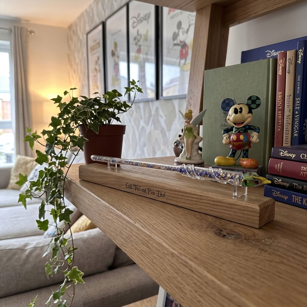 A wooden keepsake display stand engraved with 'Faith, Trust and Pixie Dust' displaying a Disney glass wand sits on a wooden shelf in a cozy living room setting, surrounded by books, a small plant, and Disney-themed figurines.