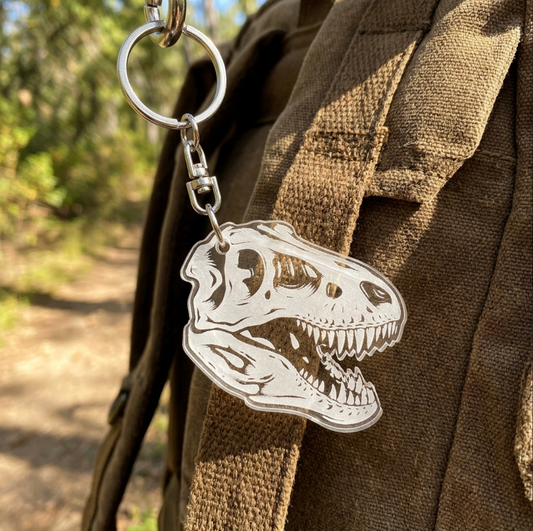 A clear acrylic T-Rex dinosaur skull keychain attached to a metal key ring and swivel clasp, shown hanging from a brown canvas backpack outdoors on a blurred forest path.