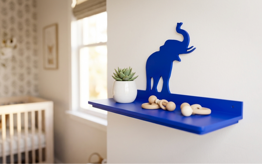 A bright blue wall-mounted elephant shelf in a nursery setting, styled with a small potted succulent and wooden rings, with a crib and window blurred in the background.
