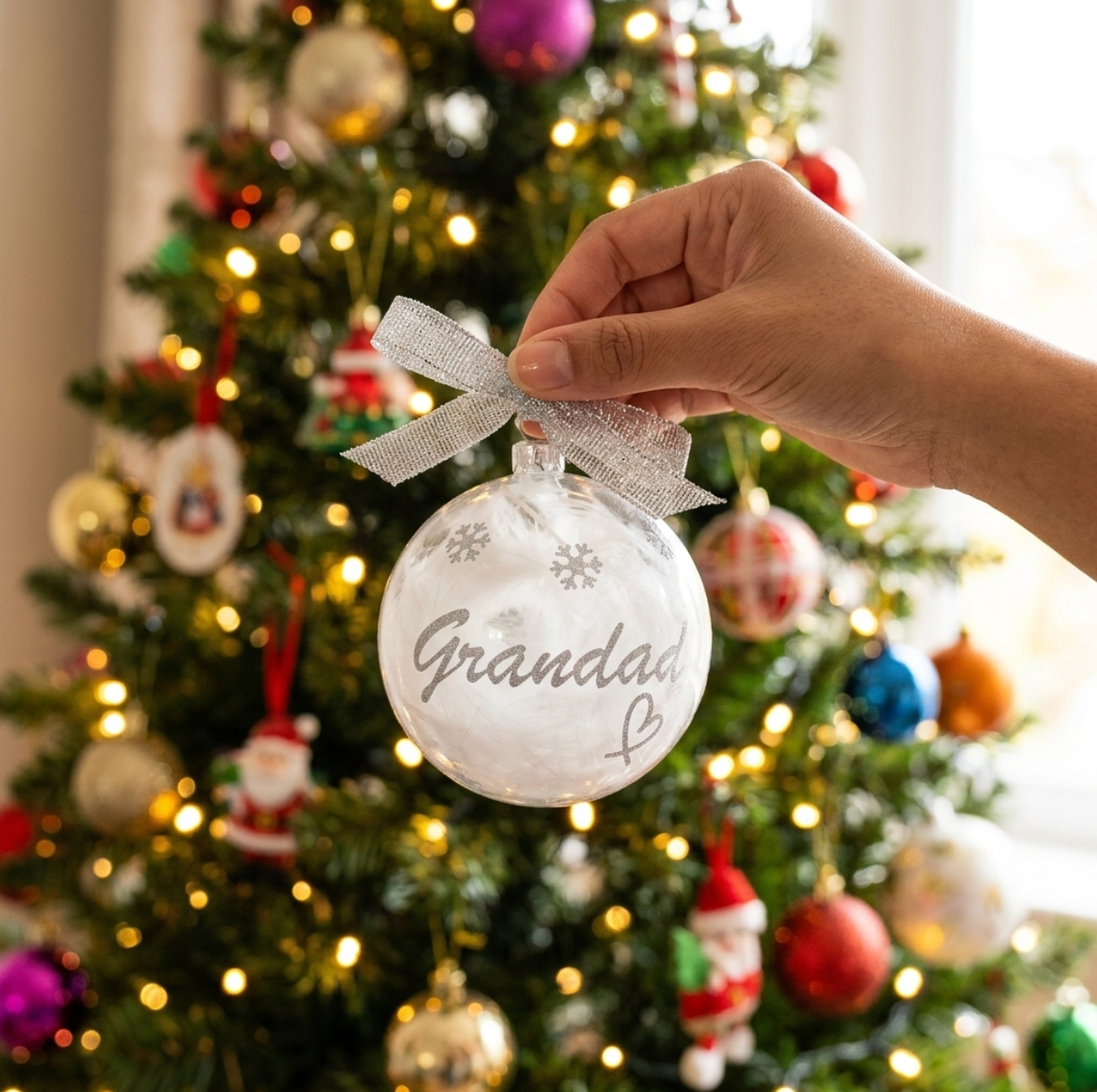A hand holds a shatterproof bauble Christmas ornament filled with white feathers and 'Grandad' written in silver vinyl and a silver organza ribbon bow, set against a blurred decorated Christmas tree with warm lights.