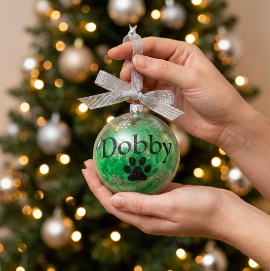 A person holds a personalised Christmas shatterproof bauble ornament filled with green feathers and the name 'Dobby' and a black paw print added, decorated with a silver glitter ribbon. A blurred Christmas tree with lights is in the background.