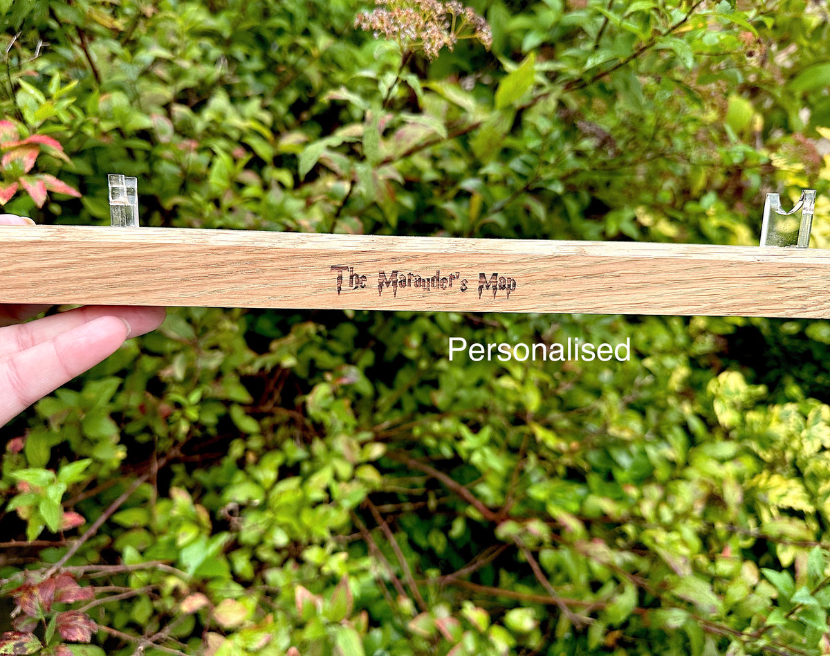 A hand holds a light-coloured wooden wand stand outdoors in front of a green leafy background. It has the words 'The Marauder's Map' engraved on it, and a white text overlay reads 'Personalised'. Harry Potter inspired wand stand.
