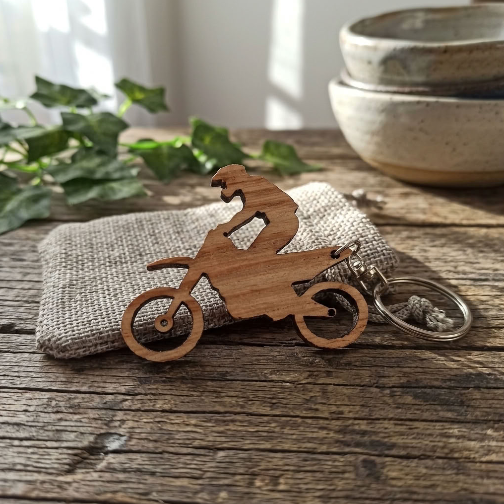 A wooden cutout keychain shaped like a motocross rider on a dirt bike, resting on a textured fabric pouch over a rustic wooden table with green leaves and ceramic bowls in the background.