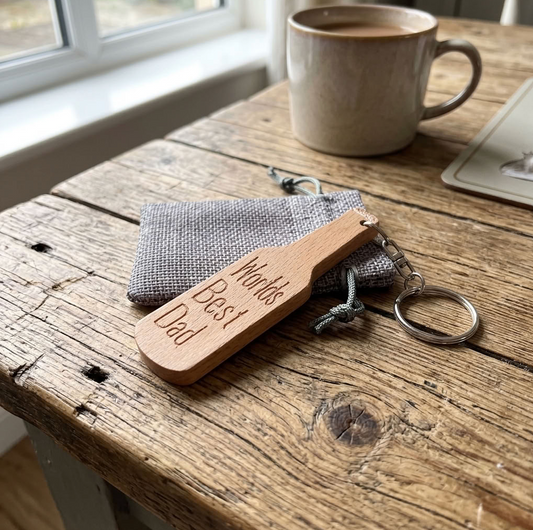 A wooden engraved keychain reading 'Worlds Best Dad' resting on a grey fabric pouch on a rustic wooden table, with a ceramic mug and a window in the background.