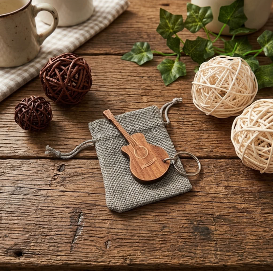 A wooden guitar-shaped keychain with a keyring, resting on a small grey fabric drawstring pouch. The scene is styled on a rustic wooden table with decorative yarn balls, green ivy, and ceramic mugs.
