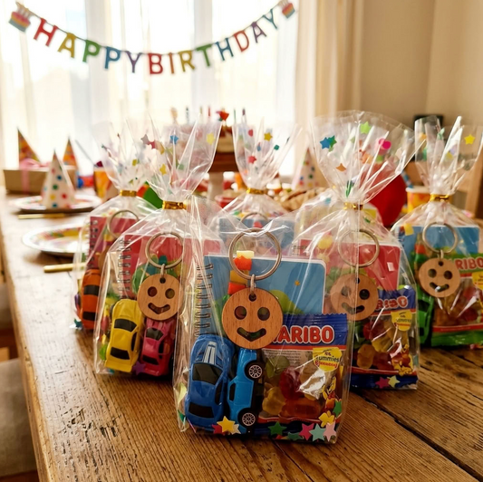 Several clear cellophane gift bags filled with birthday treats and small toy cars are arranged on a wooden table. The bags have wooden round keychains with smiley faces and a key ring at the top.