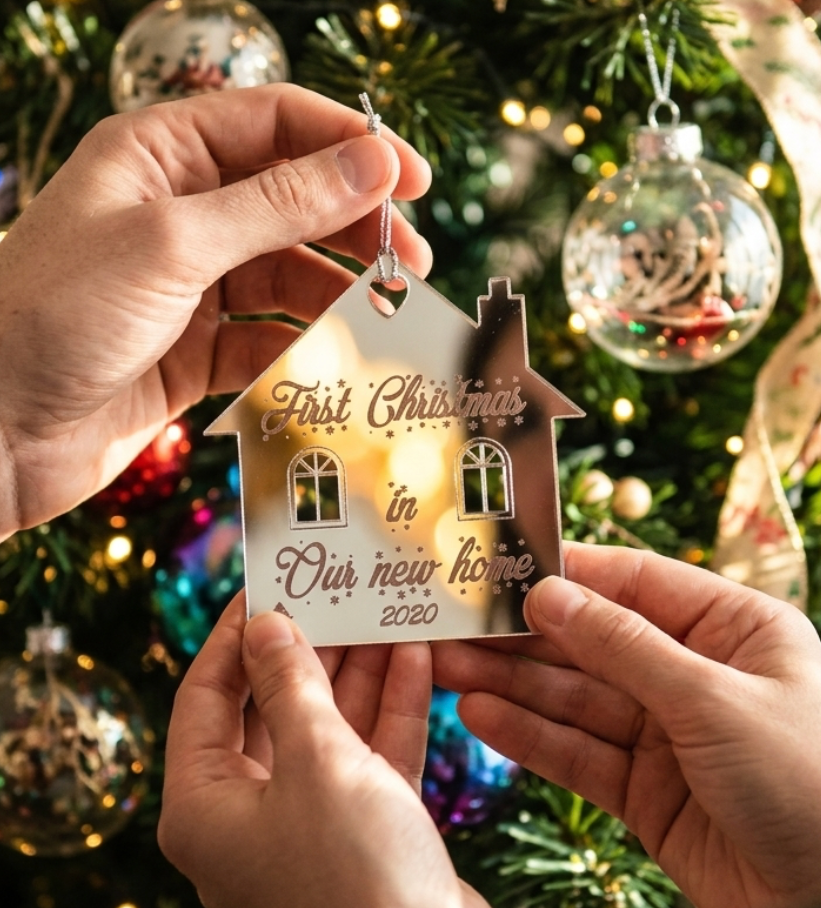 A couple holds a small house-shaped Christmas ornament with the engraved message 'First Christmas in Our new home' with the current year illuminated from within, set against a festive Christmas tree background.