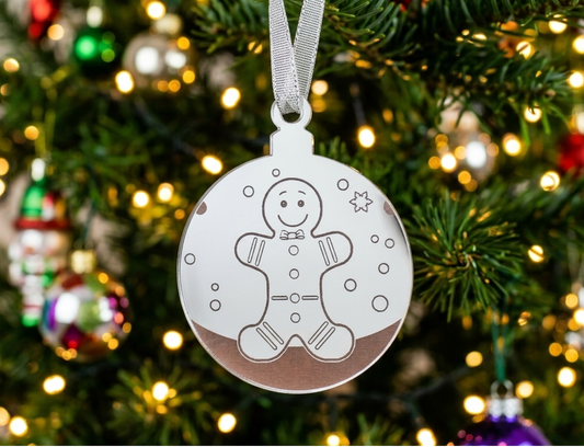 A round mirrored acrylic Christmas ornament with a gingerbread man outline and small snowflake details, hanging from a silver ribbon on a decorated Christmas tree with warm lights.
