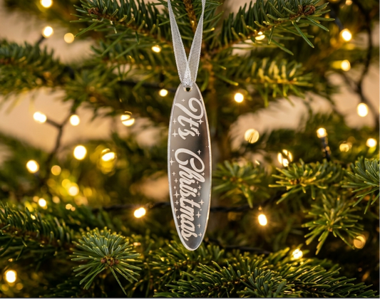 A silver mirror acrylic Christmas ornament with the words 'It's Christmas' engraved, hanging from a silver ribbon on a decorated Christmas tree with warm lights.