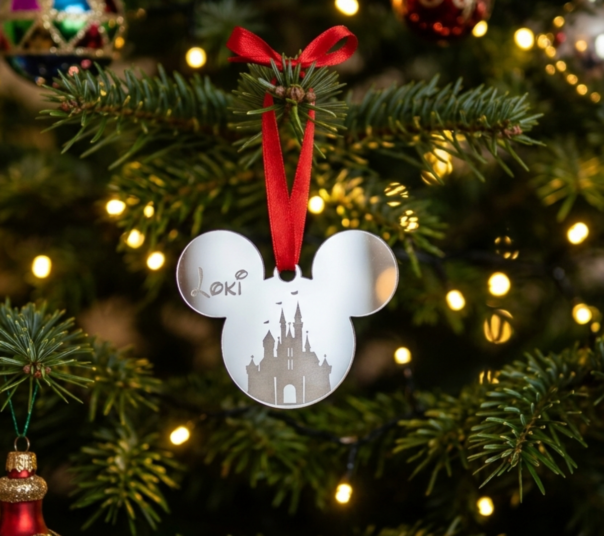 A silver mirror acrylic Mickey-shaped holiday ornament with a castle silhouette and the name 'Loki' engraved, hanging from a red ribbon on a decorated Christmas tree with warm lights.