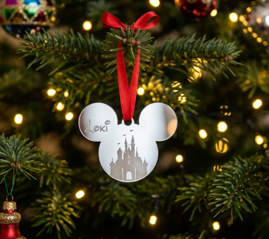 A silver mirror acrylic Mickey-shaped holiday ornament with a castle silhouette and the name 'Loki' engraved, hanging from a red ribbon on a decorated Christmas tree with warm lights.