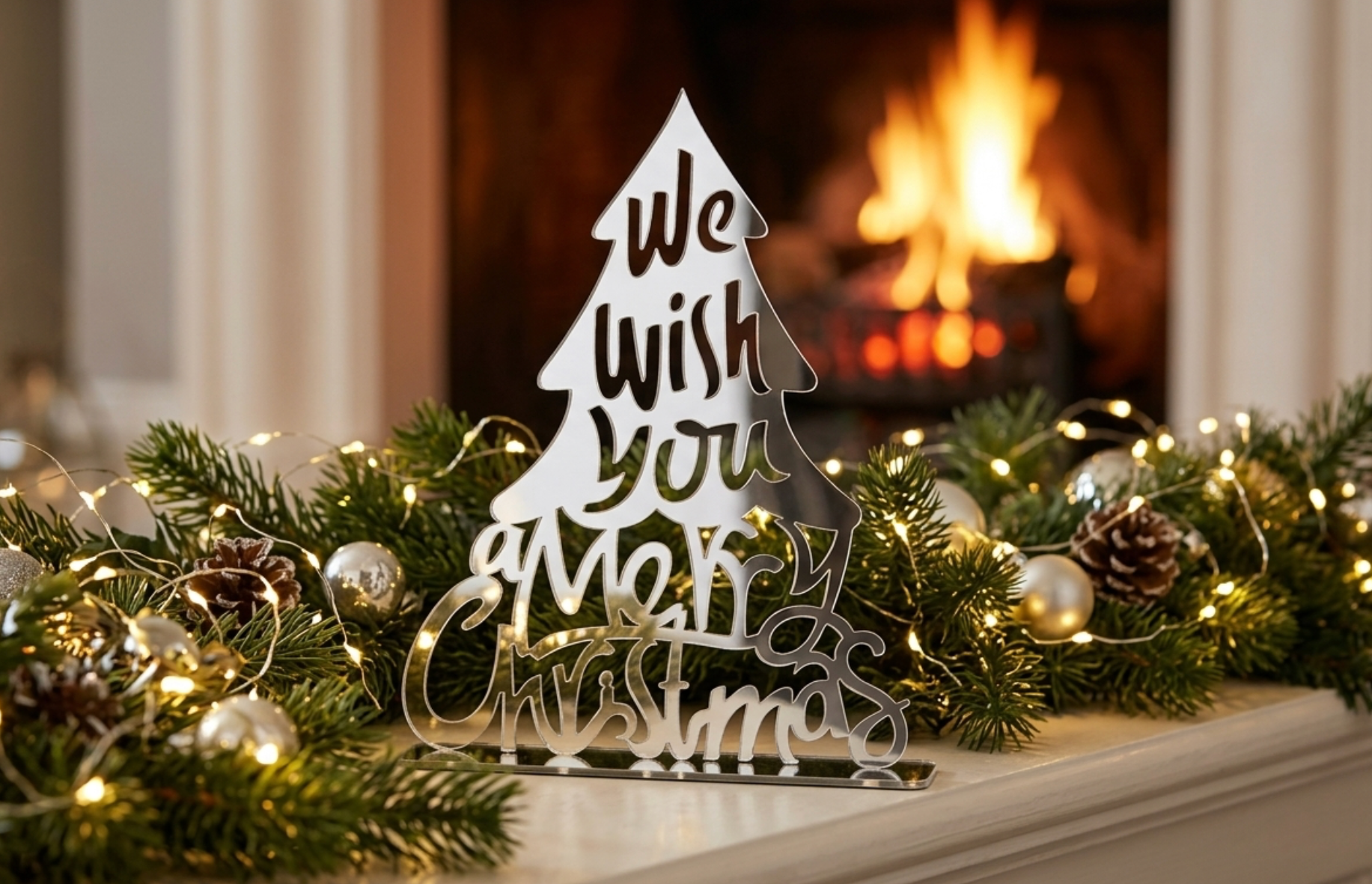A silver mirror acrylic Christmas tree sign reading 'We Wish You a Merry Christmas' styled on a fireplace mantel with a green garland, pinecones, silver and gold ornaments, and warm string lights, with a blurred fire burning in the background.