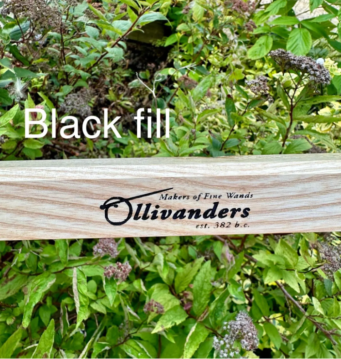 A wooden wand holder or plaque with the brand name 'Ollivanders' engraved, placed outdoors in front of dense green foliage. White text overlay reads 'Black fill'.