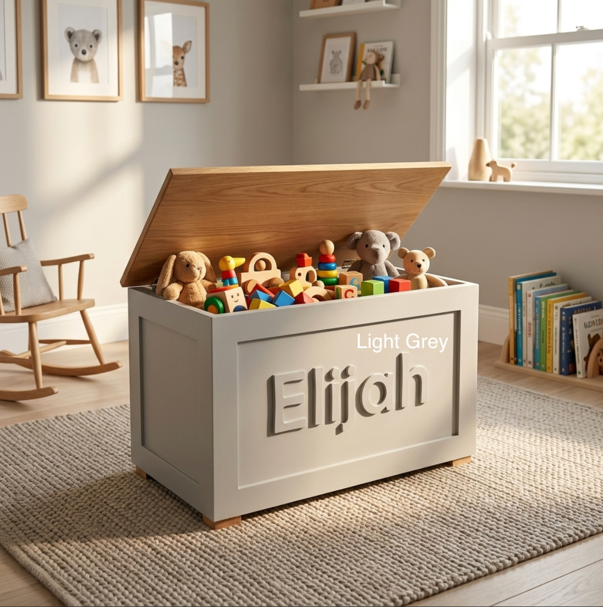 A light grey wooden toy storage chest with the name 'Elijah' cut into the front panel, shown open and filled with stuffed animals and toys in a sunlit children's room. The lid has a warm natural wood finish, and the room has a window, rocking chair, and bookshelves.