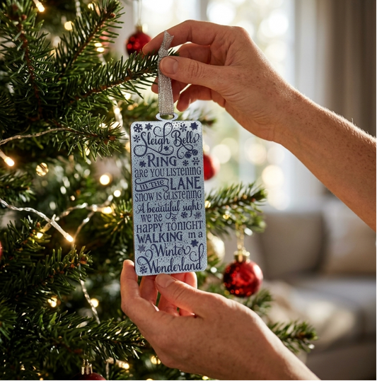 A person's hands hang a rectangular blue glitter acrylic Christmas ornament with blue text and snowflake details from a decorated Christmas tree with warm string lights and red ornaments.